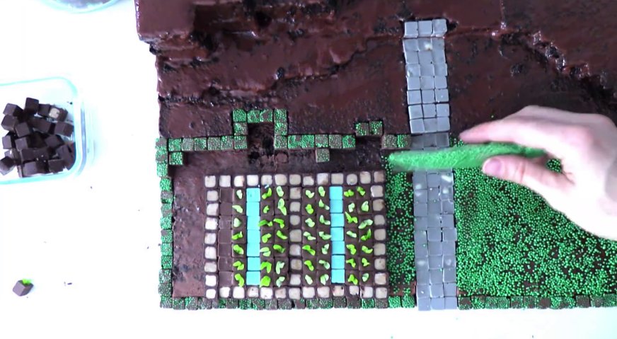 Торт майнкрафт: Заполняем поверхность бисквита ризными блоками, формируя каменные дорожки, клумбы, грядки. Промежутки посыпаем зелёными драже, формируя