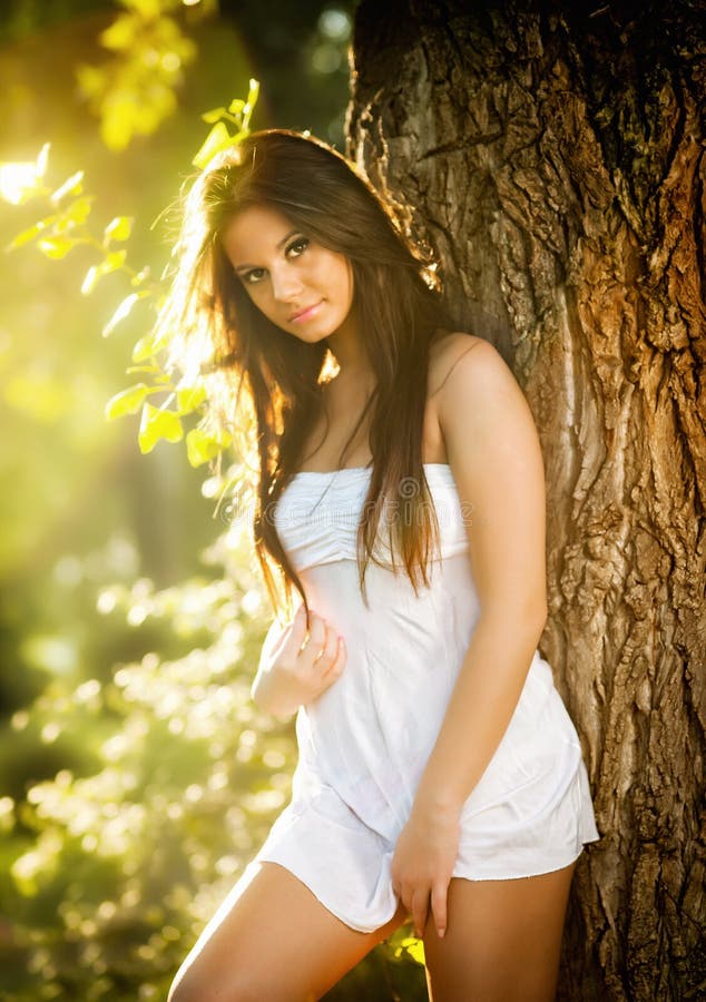 Attractive young woman in white short dress posing near a tree in a sunny summer day. Beautiful girl enjoying the nature royalty free stock image
