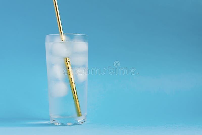 Close up of pure water with ice and gold paper straw in tall transparent glass and sun glare on blue  background. Copy space.  royalty free stock photo