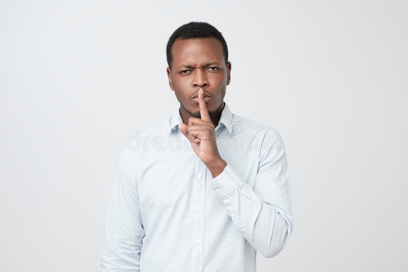 Serious african male keeps index finger on lips, asks not tell his secret to other people. Confident serious african male dressed in blue shirt, keeps index royalty free stock images