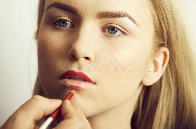 Girl getting red pencil on lips by male hand. Pretty woman with young face, makeup foundation on healthy skin and blond long hair on grey background. Visage stock photography