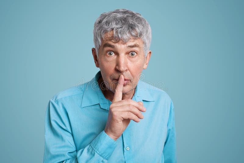 Horizontal shot of grey haired male looks with puzzled expression at camera, keeps fore finger on lips, asks to be quiet and mute,. Demands hush, dressed in royalty free stock images