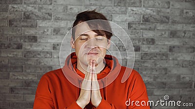 A capricious young man in a red hoodie has begging pleading expression. A male purses lips and holds hands in pray. A capricious young man in a red hoodie has stock video footage